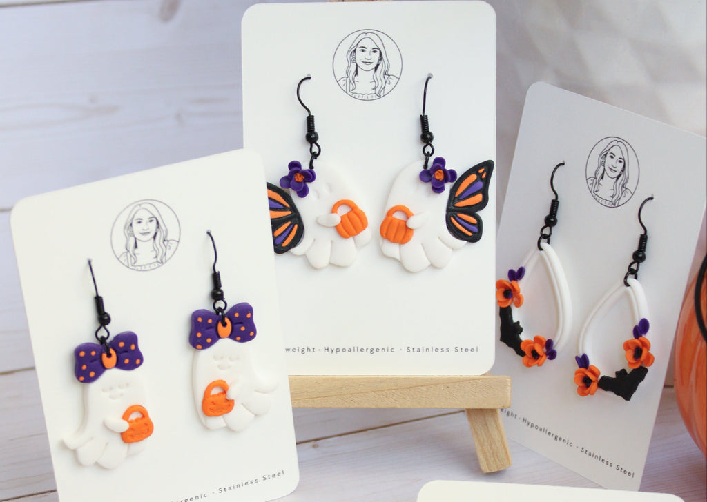 Five pairs of Halloween-themed dangle earrings displayed on white backing cards, arranged on a light wood surface. The designs include white pumpkins with orange faces, black witch hats with orange faces, purple bows with orange faces, a butterfly with purple, orange, and black details, and black flowers with gold centers.