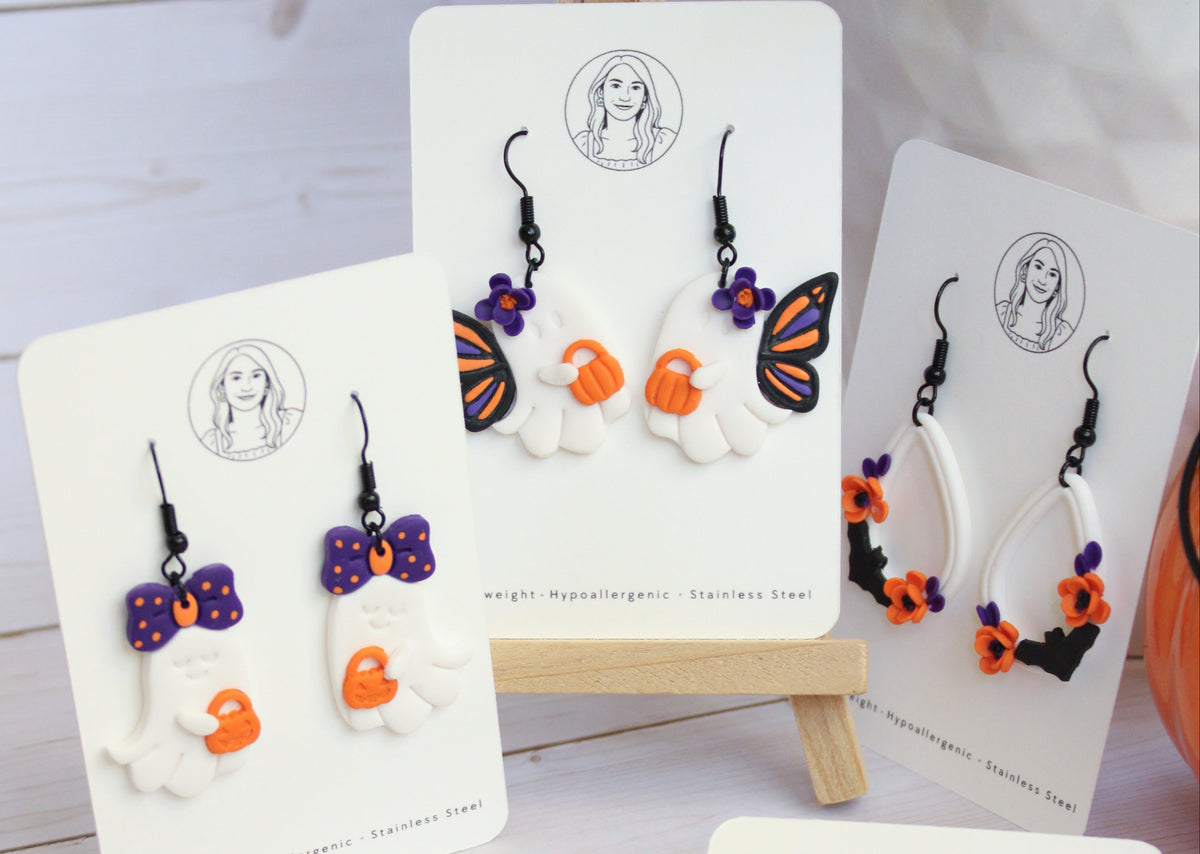 Five pairs of Halloween-themed dangle earrings displayed on white backing cards, arranged on a light wood surface. The designs include white pumpkins with orange faces, black witch hats with orange faces, purple bows with orange faces, a butterfly with purple, orange, and black details, and black flowers with gold centers.